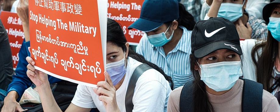 Masked women sit in protest against the military coup, holding red signs in English and Burmese. Photo credit: Wikicommons/ MgHla.
